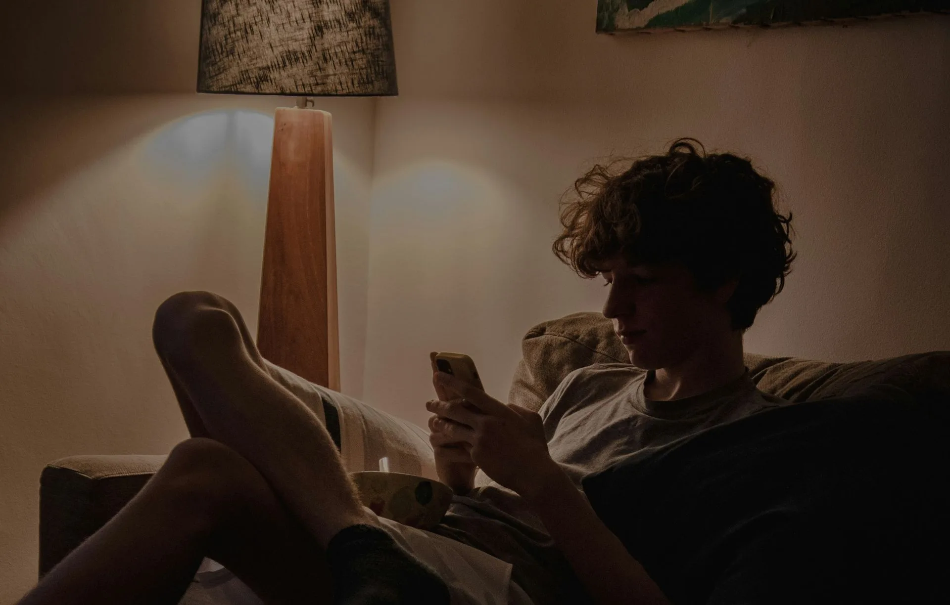 Jemand ruht abends auf dem Sofa, die Füße hochgelegt, das Telefon in der Hand, im warmen Licht einer Stehlampe