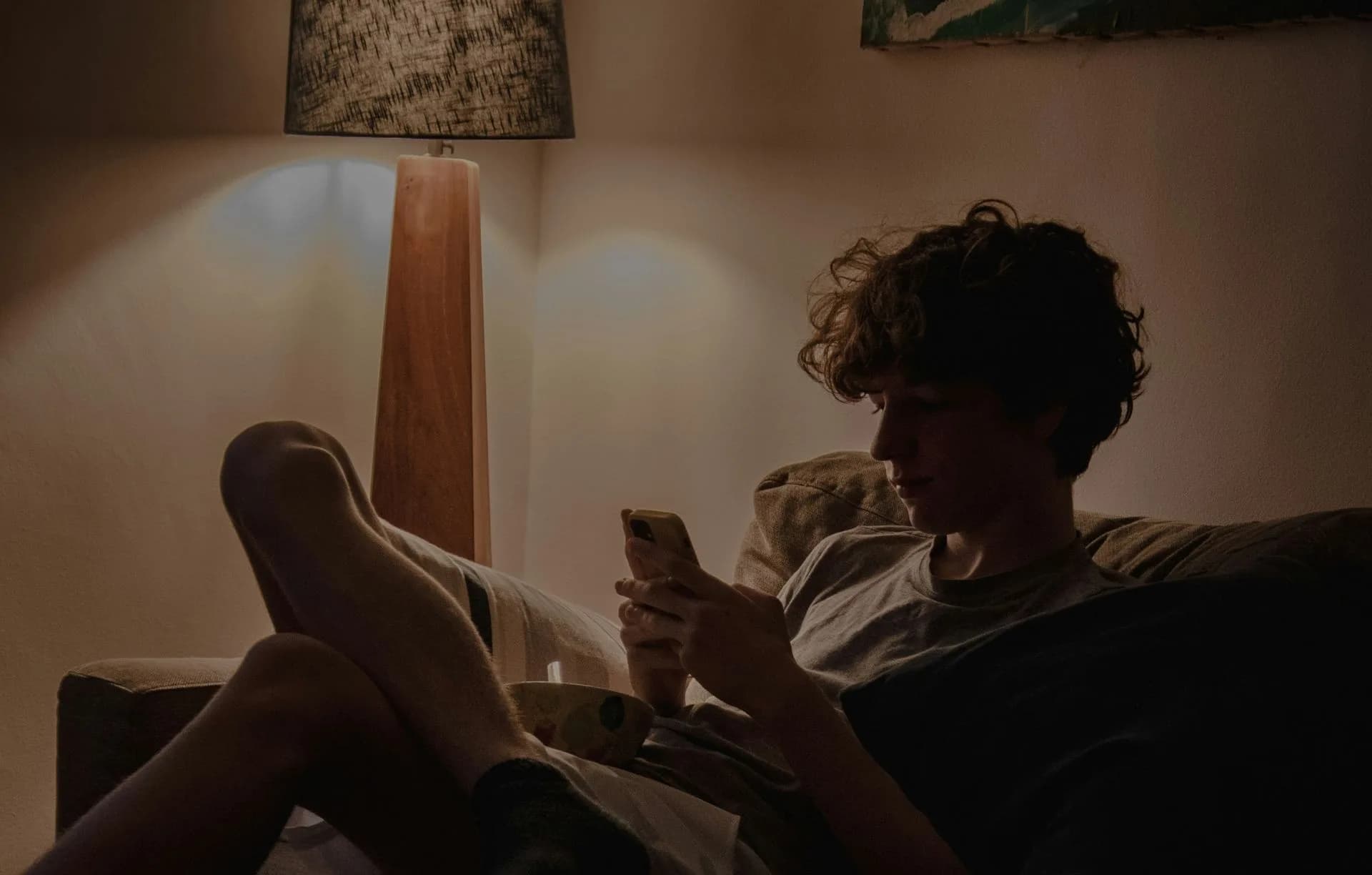 Jemand ruht abends auf dem Sofa, die Füße hochgelegt, das Telefon in der Hand, im warmen Licht einer Stehlampe